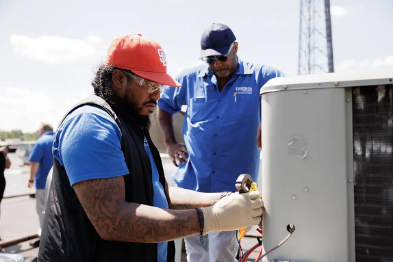 An HVAC student working on an AC unit with an instructor watching